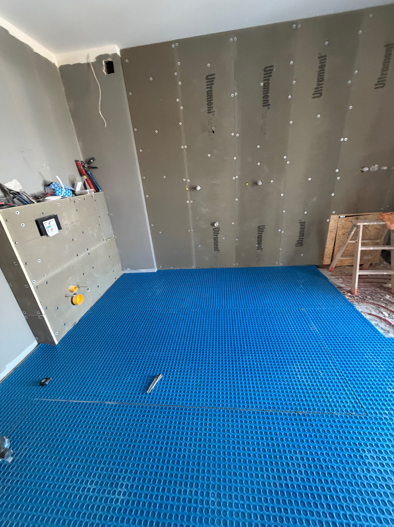 Niebieska mata pod ogrzewanie podłogowe w trakcie montażu, ściany wyłożone płytami Ultrament, drabina w tle. Etap prac przed wylaniem posadzki.