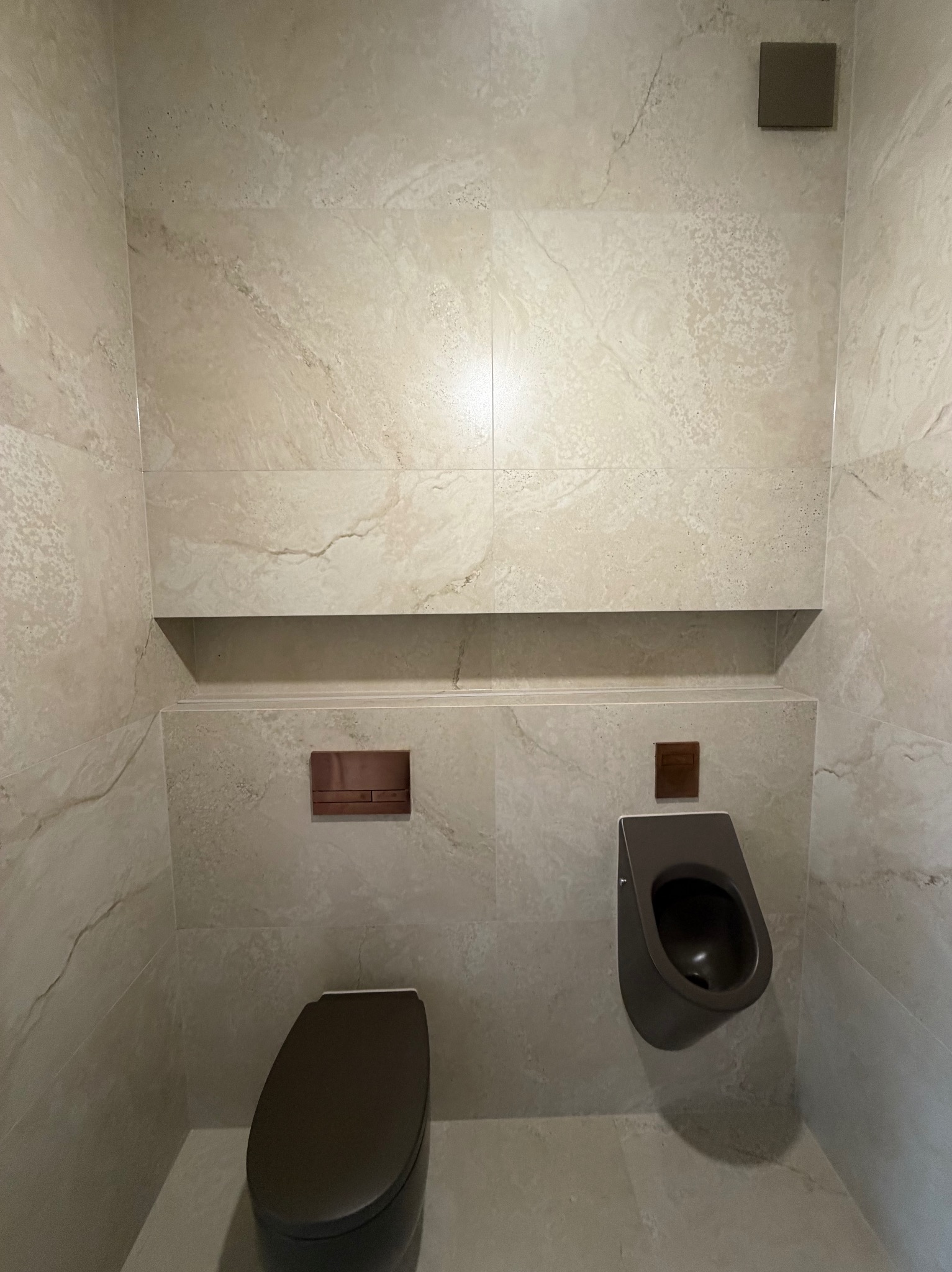 Nowoczesna łazienka z beżowymi płytkami imitującymi marmur, wiszącą toaletą i pisuaru w kolorze grafitowym. Minimalistyczny design z wnęką i ukrytymi przyciskami.