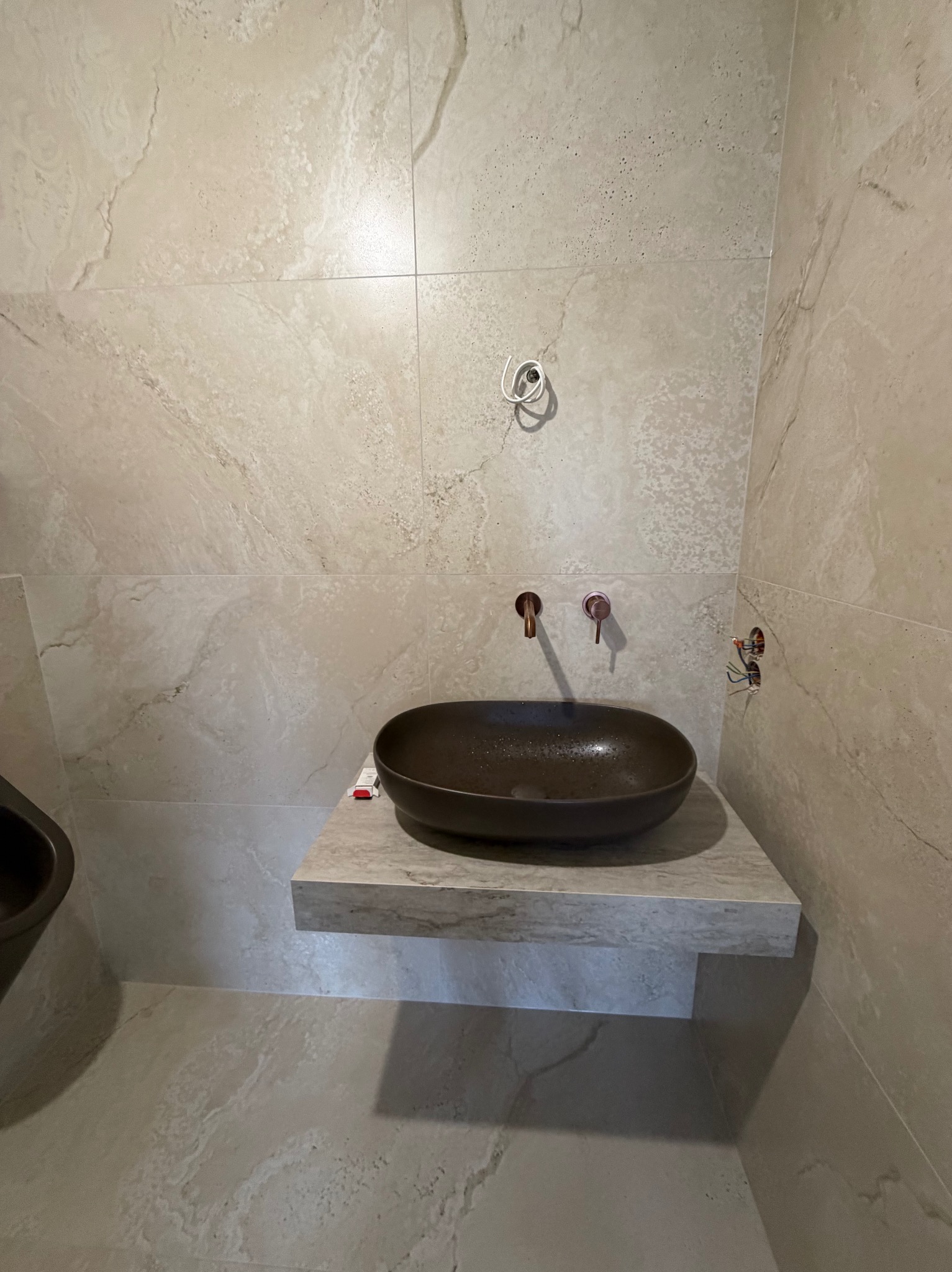 Beżowa łazienka z owalną, ciemną umywalką na kamiennym blacie. Widoczne miedziane krany i fragment instalacji elektrycznej. Płytki imitujące kamień na ścianach i podłodze.