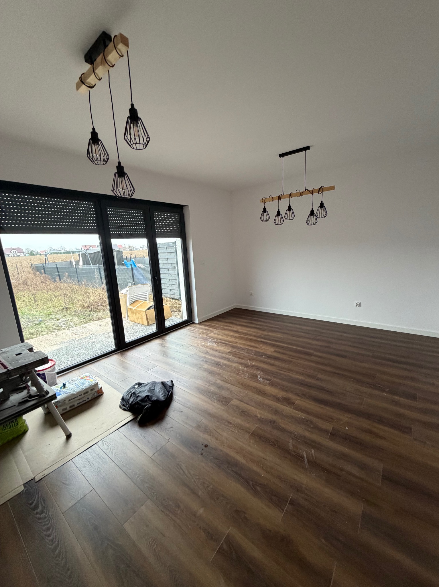 Jasne wnętrze z panelami podłogowymi i nowoczesnym oświetleniem. Widoczne okno z roletami zewnętrznymi i elementy przygotowania do prac wykończeniowych.