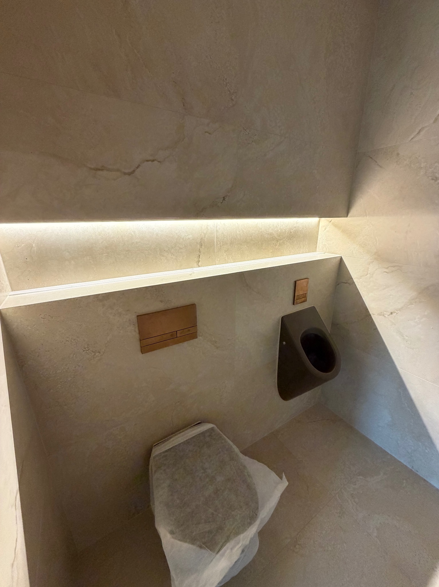 Nowoczesna toaleta w beżowych płytkach z podświetleniem LED, wiszącym bidetem i ukrytą spłuczką. Częściowo zakryta miska WC folią ochronną.