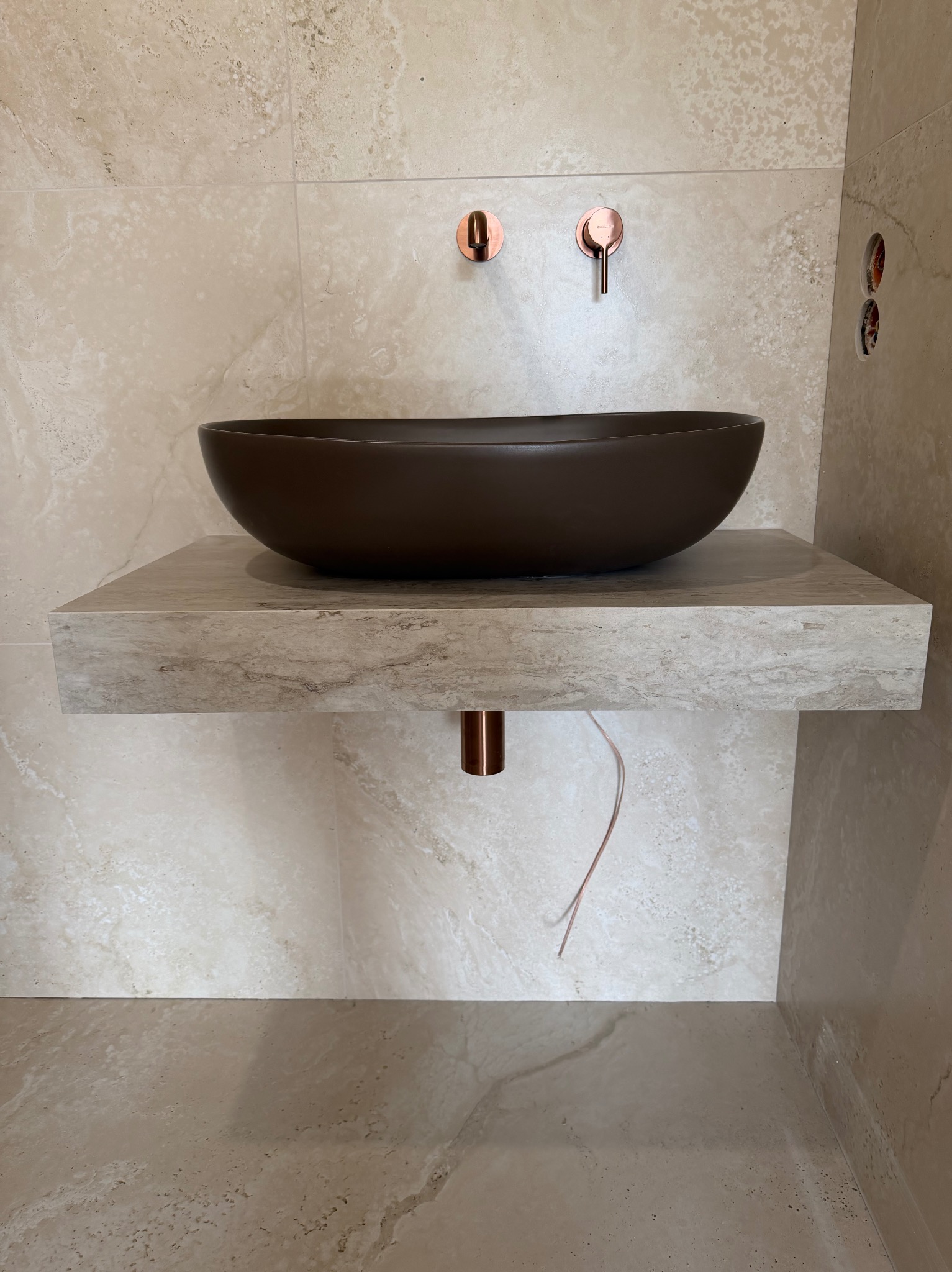 Beżowa łazienka z umywalką w kształcie misy na kamiennym blacie, miedziana armatura i płytki imitujące trawertyn. Minimalistyczny design z widocznymi przewodami.
