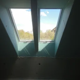 Montaż płyty karton gips na poddaszu z dwoma oknami dachowymi, widok na zewnątrz z zielenią i niebem.