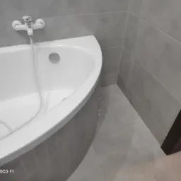 Biała, narożna wanna z chromowaną armaturą na tle szarych płytek ściennych i podłogowych, widoczny fragment ciemnobrązowej framugi drzwi.