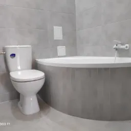 Jasna łazienka z szarymi płytkami: widok na toaletę Cersanit, okrągłą wannę z baterią i dwa małe, kwadratowe włazy rewizyjne na ścianie.