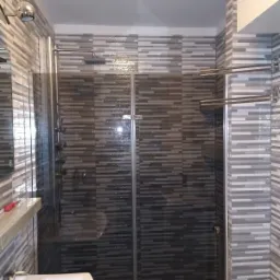 Kabina prysznicowa z ciemnymi płytkami w paski, widoczny panel prysznicowy z hydromasażem, umywalka z baterią i grzejnik drabinkowy.