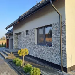 Elewacja domu wykończona kamieniem dekoracyjnym, z widocznym oknem, rynną i fragmentem chodnika z kostki brukowej. Widoczne nasadzenia krzewów ozdobnych.