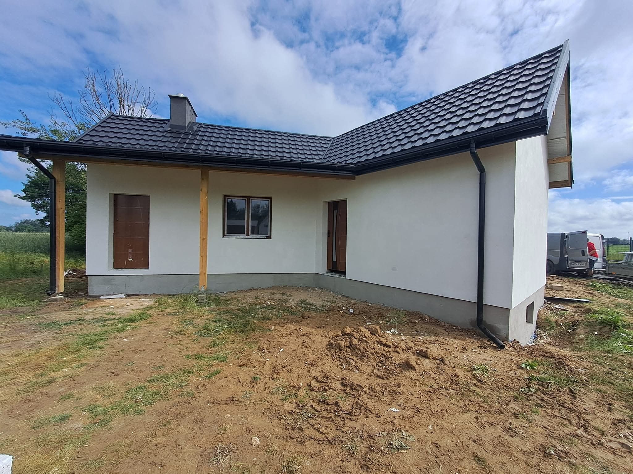 Nowoczesny dom jednorodzinny z ciemną dachówką i białymi ścianami, widok z zewnątrz na tle błękitnego nieba z chmurami. Widoczne elementy wykończeniowe i teren wokół domu.