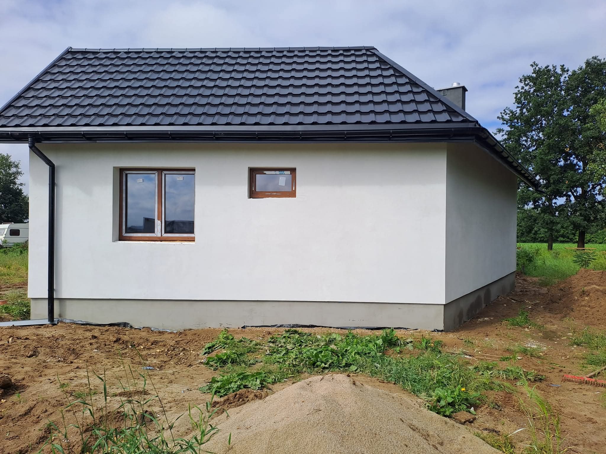 Boczny widok domu z białą elewacją, ciemnym dachem i brązowymi oknami. W tle zieleń i błękitne niebo. U dołu widoczny piasek i roślinność.