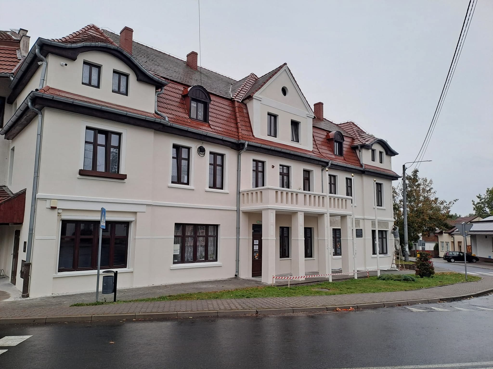 Odnowiona fasada budynku z czerwonym dachem i ciemnymi oknami, widoczna z ulicy po deszczu. Świeżo pomalowane ściany w jasnym odcieniu, schludny wygląd.