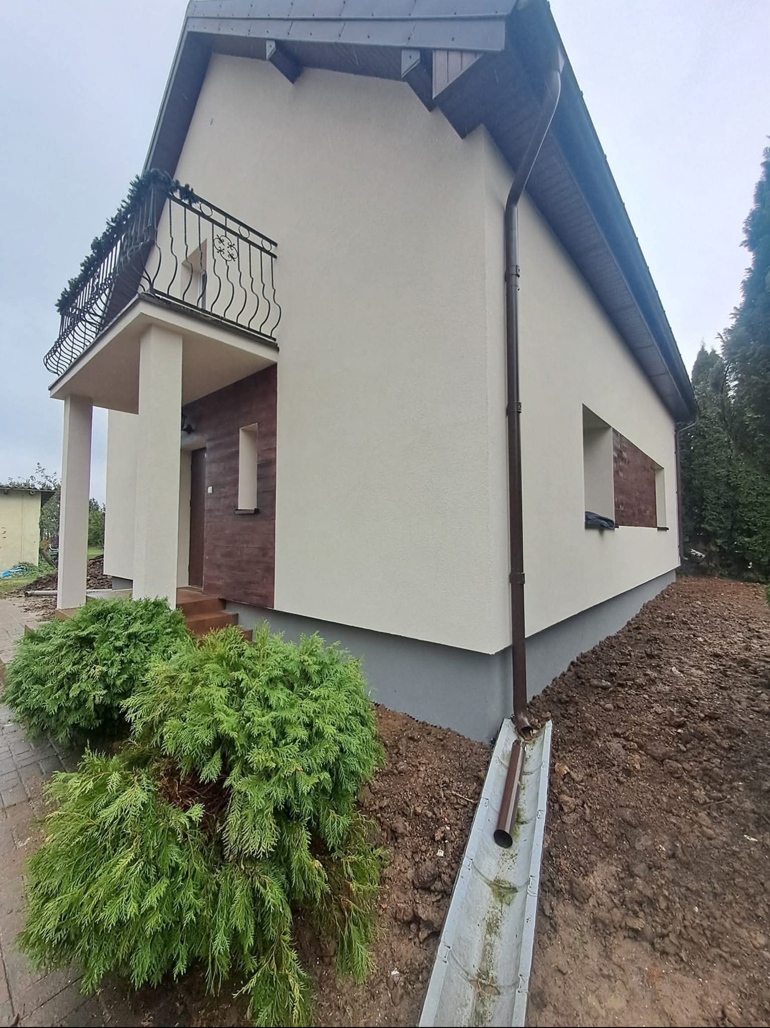 Elewacja domu po malowaniu w jasnym odcieniu, z dekoracyjnym balkonem i brązowymi elementami wykończeniowymi. Widoczne krzewy i fragment chodnika.