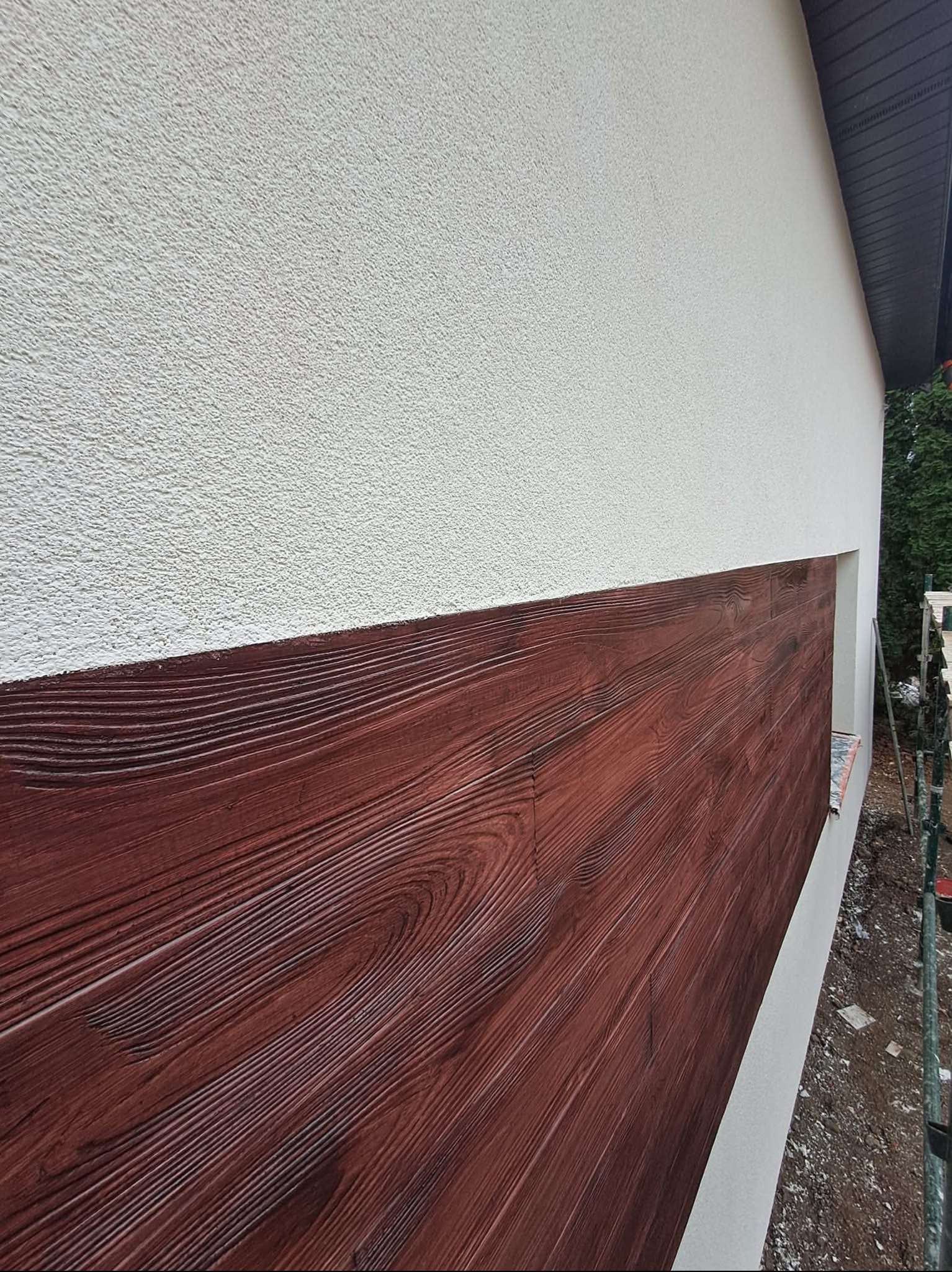 Fragment elewacji domu z dekoracyjnym panelem imitującym drewno, obok okna, widoczne rusztowanie. Górna część ściany pokryta jasnym tynkiem.