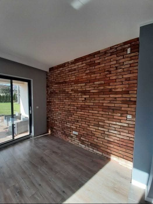 Ściana wykończona cegłą dekoracyjną w odcieniach brązu i czerwieni, widok na parkiet i okno przesuwne. Nowoczesne wnętrze po remoncie.