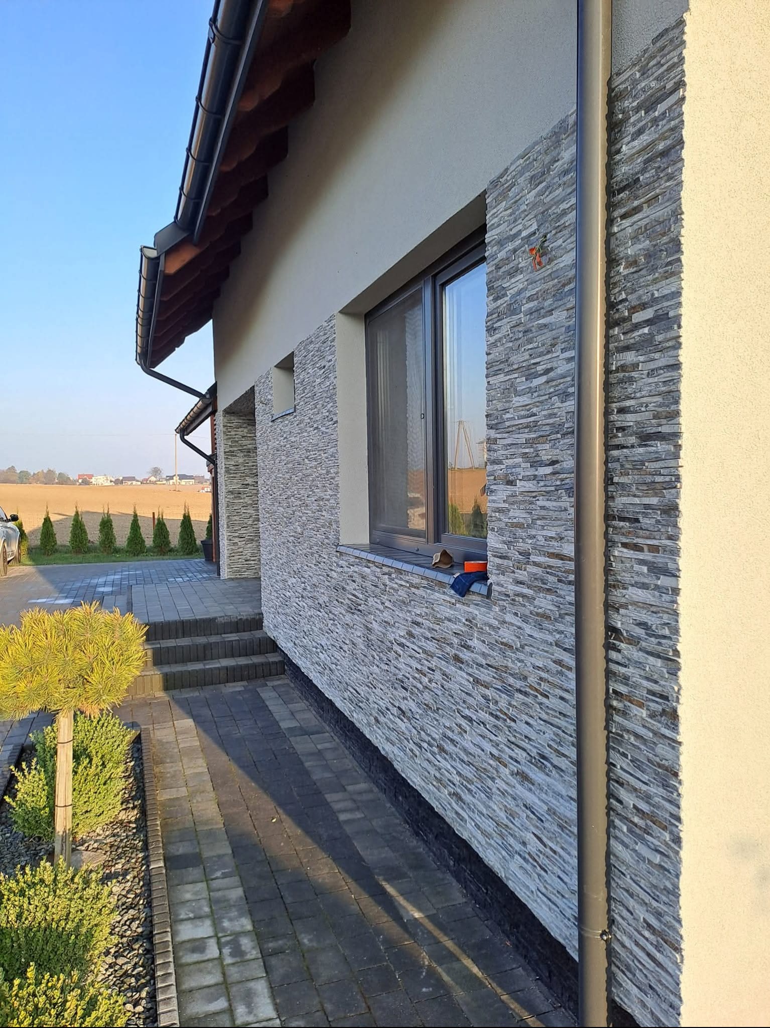 Elewacja domu z dekoracyjnym kamieniem, widok z boku na okno, rynnę i chodnik z kostki brukowej. W tle widoczne drzewa i pole. Słoneczny dzień.