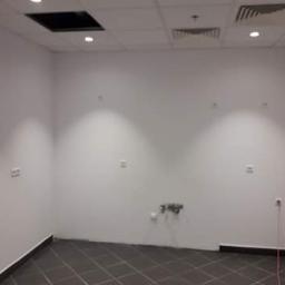 ATE Uslugi budowlane - Puste, pomalowane na biało pomieszczenie biurowe z ciemną podłogą w kratkę, widoczne gniazdka elektryczne, otwór w suficie i kratki wentylacyjne.