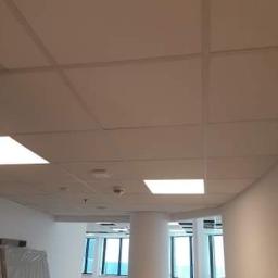 ATE Uslugi budowlane - Kasetonowy sufit podwieszany z wbudowanymi panelami LED w jasnym biurze, widok z dołu, ściany w kolorze złamanej bieli, kolumna nośna w tle.