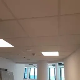 Kasetonowy sufit podwieszany z wbudowanymi panelami LED w jasnym biurze, widok z dołu, ściany w kolorze złamanej bieli, kolumna nośna w tle.