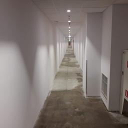 ATE Uslugi budowlane - Długi, pusty korytarz z białymi ścianami i szarą posadzką w trakcie prac wykończeniowych, widoczne nierówności i ślady po szpachlowaniu na podłodze, oświetlenie sufitowe punktowe, w tle drzwi...