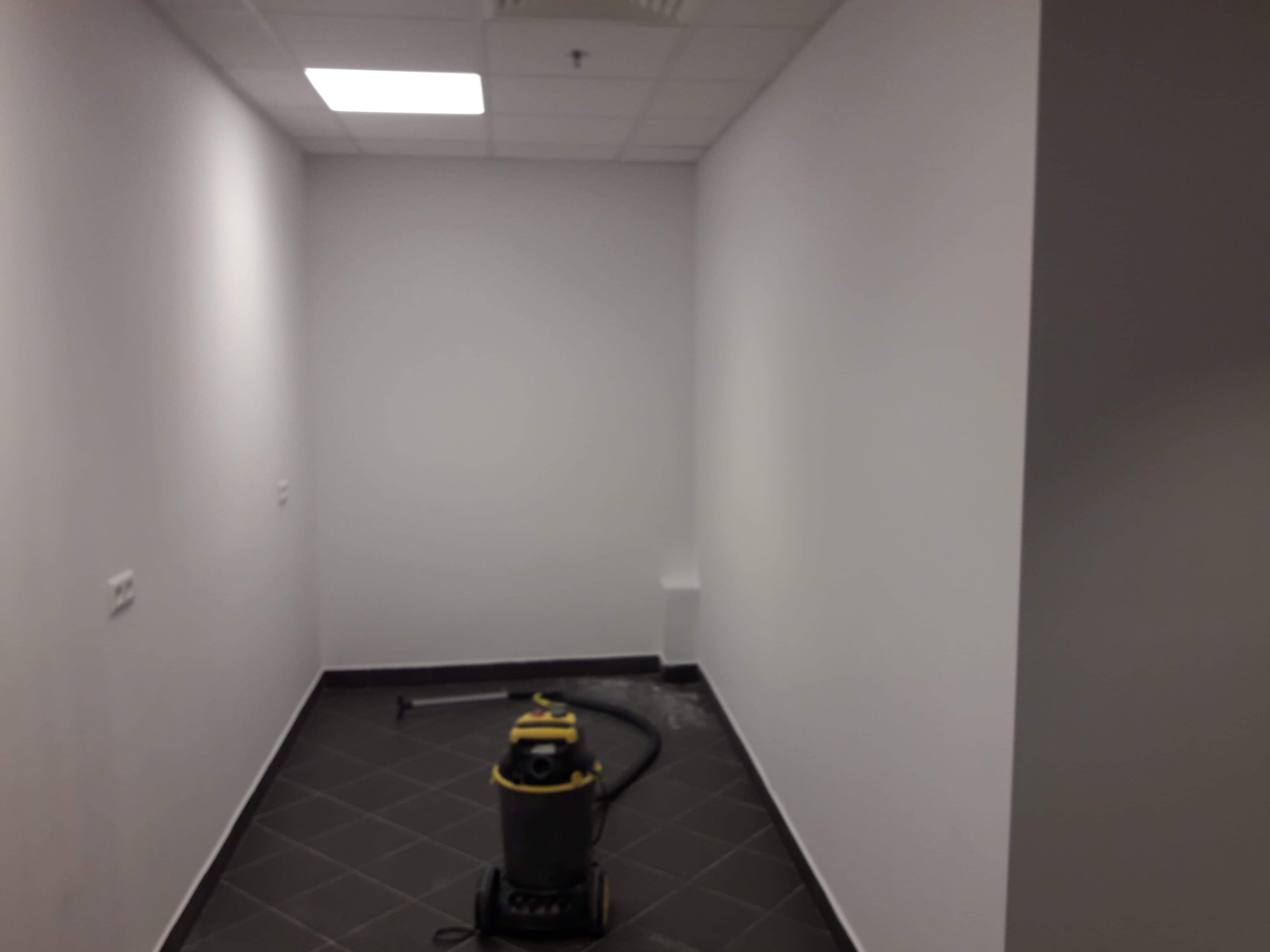 Wnętrze biurowe z szarą podłogą w romby i białymi ścianami, z widocznym odkurzaczem przemysłowym w rogu, sugerujące sprzątanie po remoncie lub regularne czyszczenie.