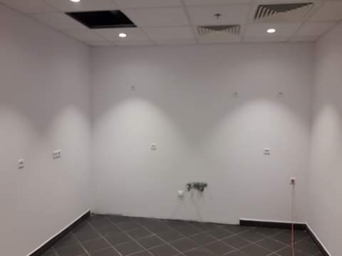 Puste, pomalowane na biało pomieszczenie biurowe z ciemną podłogą w kratkę, widoczne gniazdka elektryczne, otwór w suficie i kratki wentylacyjne.