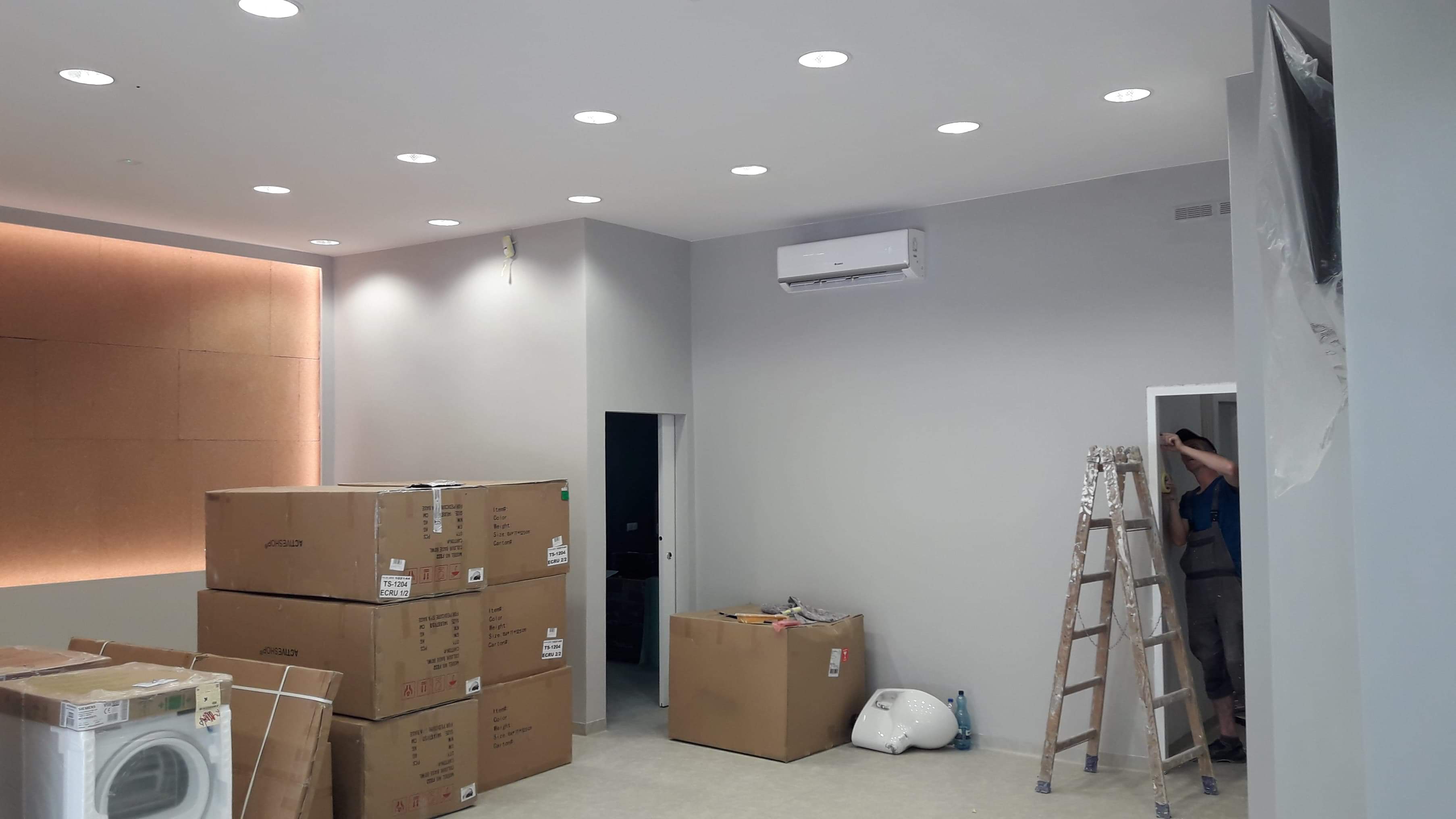 Wnętrze w trakcie prac wykończeniowych: kartony z wyposażeniem, drabina, klimatyzator na ścianie, oświetlenie sufitowe, pracownik mierzący otwór drzwiowy.