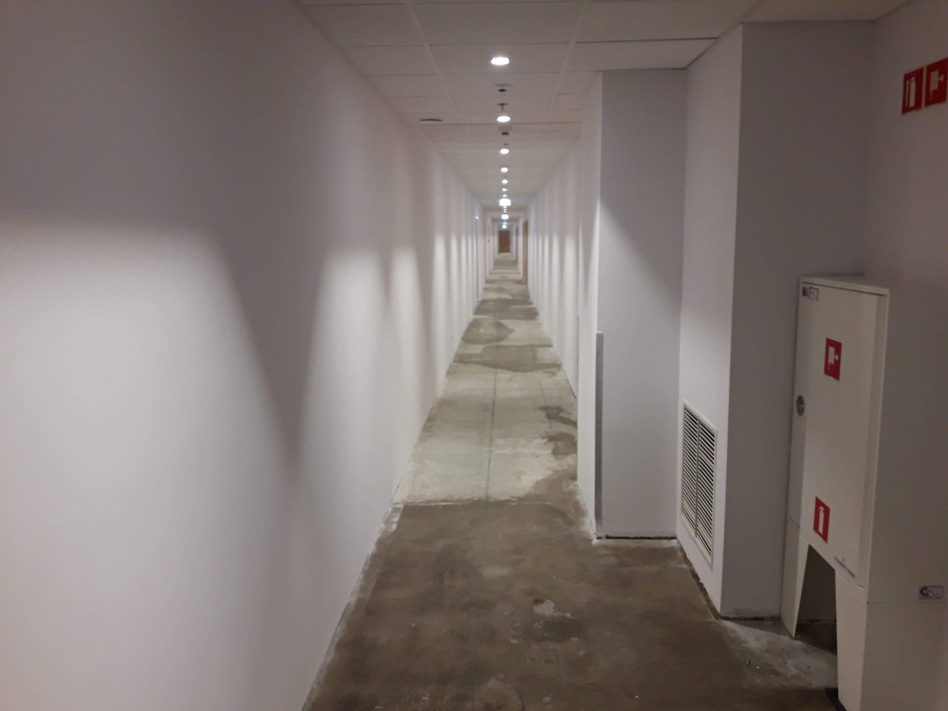 Długi, pusty korytarz z białymi ścianami i szarą posadzką w trakcie prac wykończeniowych, widoczne nierówności i ślady po szpachlowaniu na podłodze, oświetlenie sufitowe punktowe, w tle drzwi...