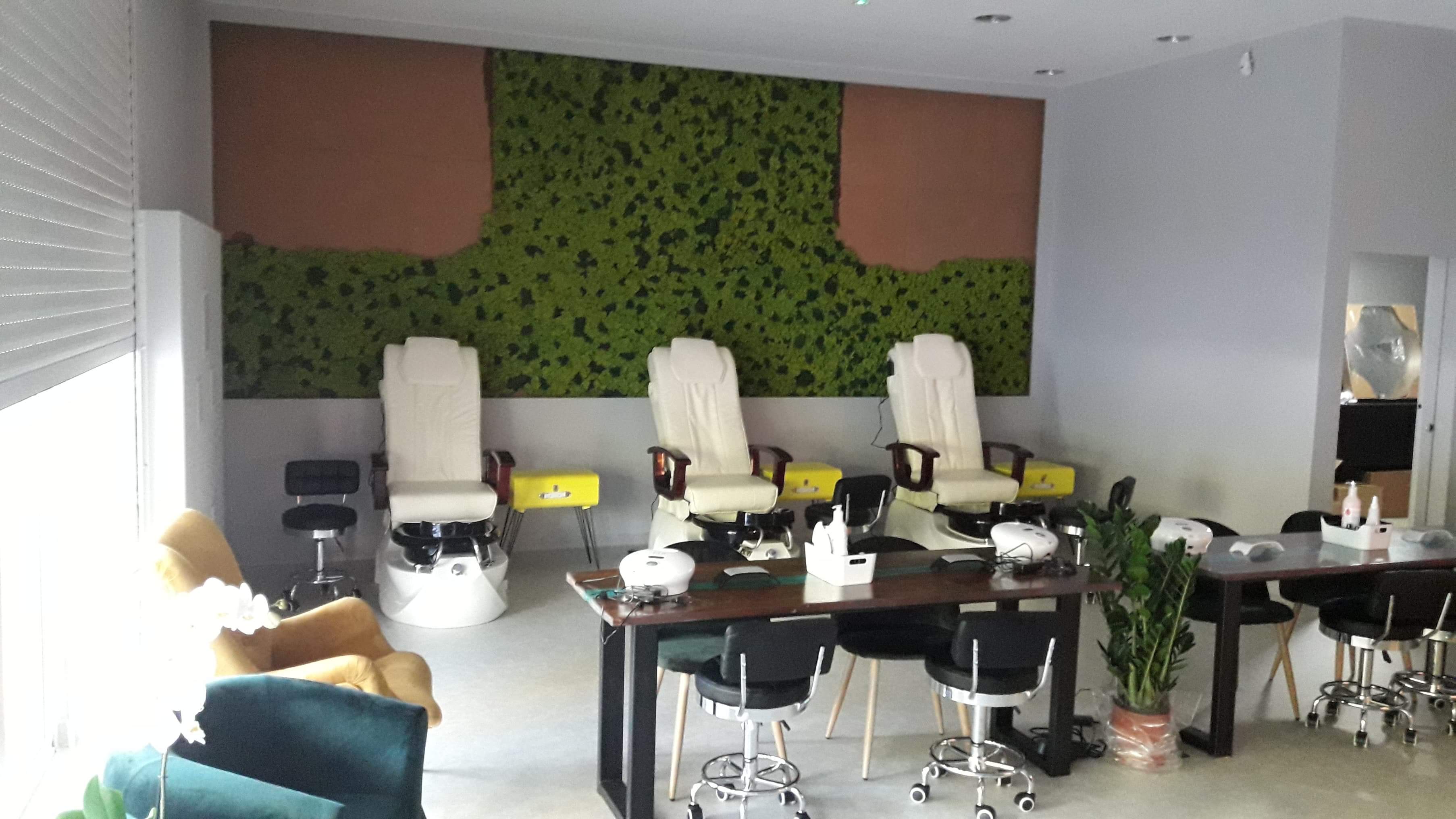 Jasne wnętrze salonu kosmetycznego z trzema fotelami do pedicure, stolikami do manicure, ścianą z mchu i brązowymi panelami, oraz elementami dekoracyjnymi.