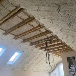 Poddasze z widoczną konstrukcją drewnianą pokryte grubą warstwą białej piany izolacyjnej, okna dachowe wpuszczające światło, zwisające przewody elektryczne.