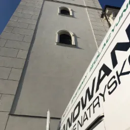 Boczny widok na wysoką, jasną kamienicę z oknami łukowymi, częściowo zasłoniętą przez białą furgonetkę z czarnym napisem reklamującym systemy izolacyjne.