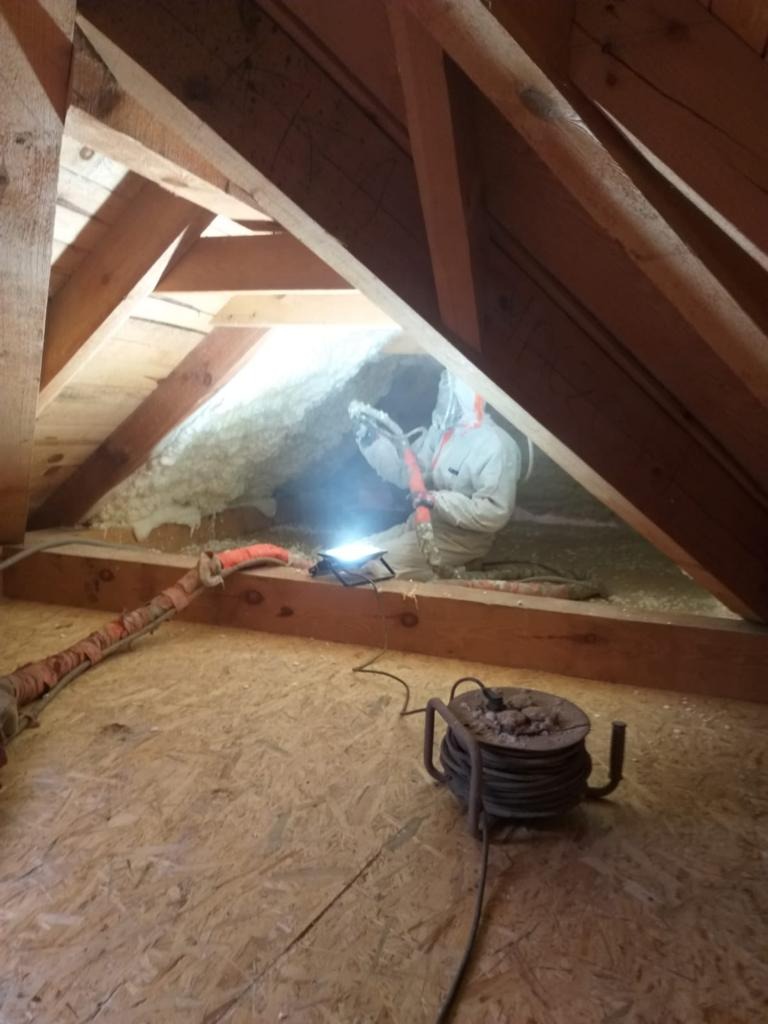 Pracownik w kombinezonie ochronnym aplikuje izolację natryskową w przestrzeni poddasza, widoczne drewniane krokwie i sprzęt do natrysku.