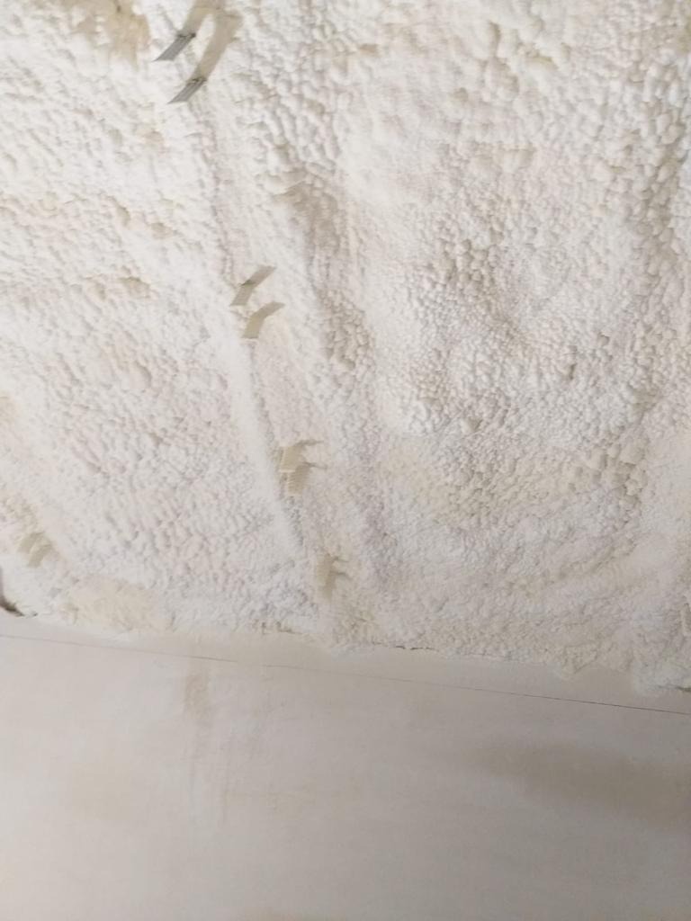 Świeżo natryśnięta, biała izolacja piankowa na suficie, widoczne metalowe elementy montażowe, dolna część ściany pomalowana na biało.