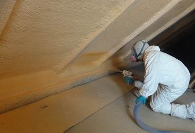 Pracownik w kombinezonie ochronnym aplikuje pianę izolacyjną na skośną powierzchnię poddasza, widoczne świeżo nałożona warstwa piany i przewód doprowadzający materiał.