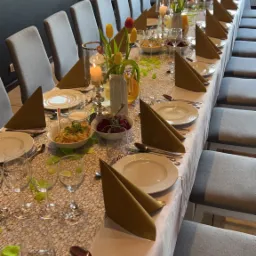 Długi stół nakryty do uroczystego posiłku z potrawami, kwiatami i świecami, ustawiony wzdłuż rzędu szarych krzeseł, z widocznymi elementami dekoracyjnymi w postaci zielonych listków.