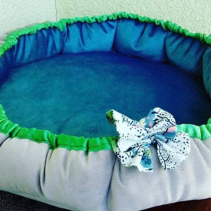 Owalne legowisko dla zwierząt w odcieniach błękitu, szarości i zieleni, ozdobione białą kokardką w czarne i niebieskie wzory.