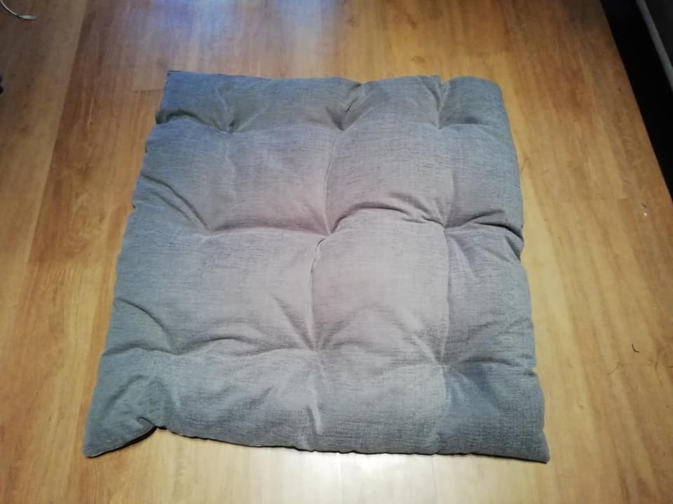 Szara, kwadratowa poduszka siedziskowa pikowana w dziewięć kwadratów, leżąca na drewnianej podłodze.