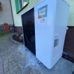 Zewnętrzna jednostka pompy ciepła Mitsubishi Electric Zubadan R32 zamontowana na betonowej podstawie przy budynku z zielonymi ramami okiennymi i brukowanym chodnikiem.