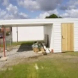 Biała, nowoczesna wiata garażowa z pomieszczeniem gospodarczym w trakcie budowy. Widoczne materiały budowlane i rusztowanie. Tło stanowi trawnik i częściowo widoczny budynek.