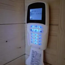 Biały panel sterowania alarmem z podświetlanymi na niebiesko przyciskami, zamontowany na drewnianej ścianie, z otwartą klapką instrukcji.