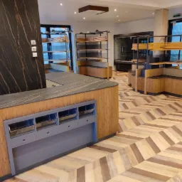 Wnętrze sklepu z blatem z ciemnego kamienia i drewnianymi regałami na podłodze w jodełkę, widoczna ściana z marmuru.