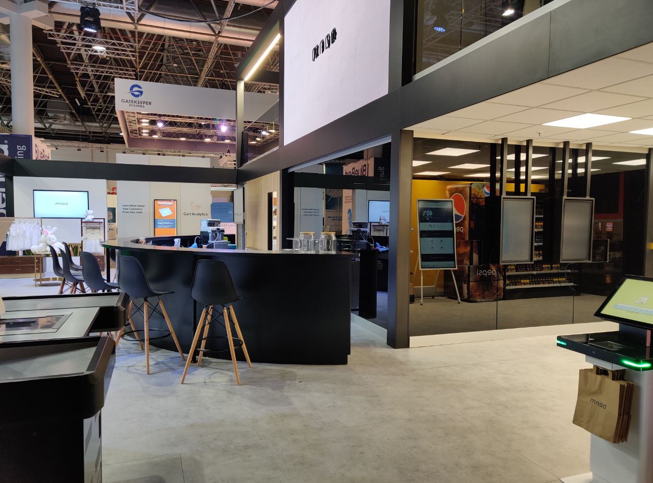 Widok stoiska targowego z czarnym barem i krzesłami na drewnianych nogach, w tle ekspozycja produktów i firmowe logo na ekranie.