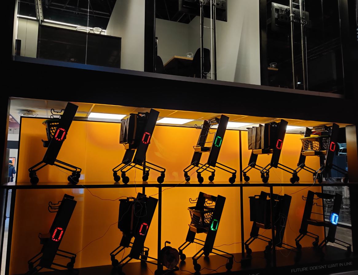 Rząd czarnych wózków sklepowych z wyświetlaczami LED prezentowanych na pomarańczowym tle w nowoczesnej przestrzeni wystawienniczej.