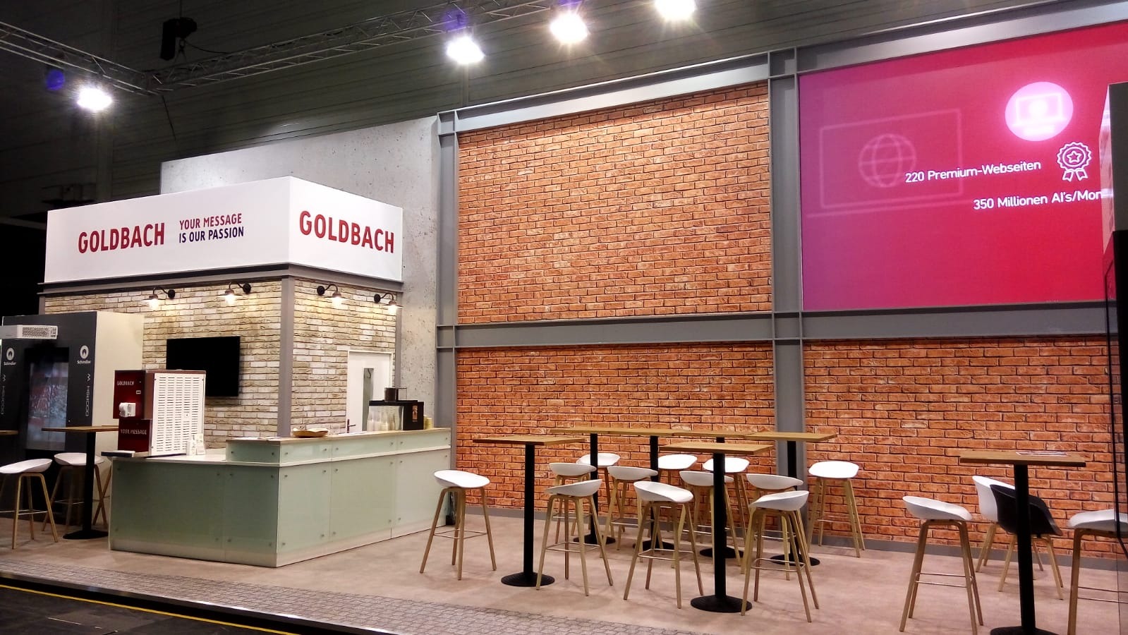 Stoisko targowe firmy Goldbach z ceglaną ścianą w tle, wyposażone w wysokie stoliki barowe i białe hokery, z podświetlanym panelem reklamowym prezentującym statystyki internetowe.