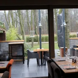 Zadaszony taras restauracji z widokiem na ogród, stoliki nakryte do posiłku, na zewnątrz widoczne drzewa i elementy małej architektury.