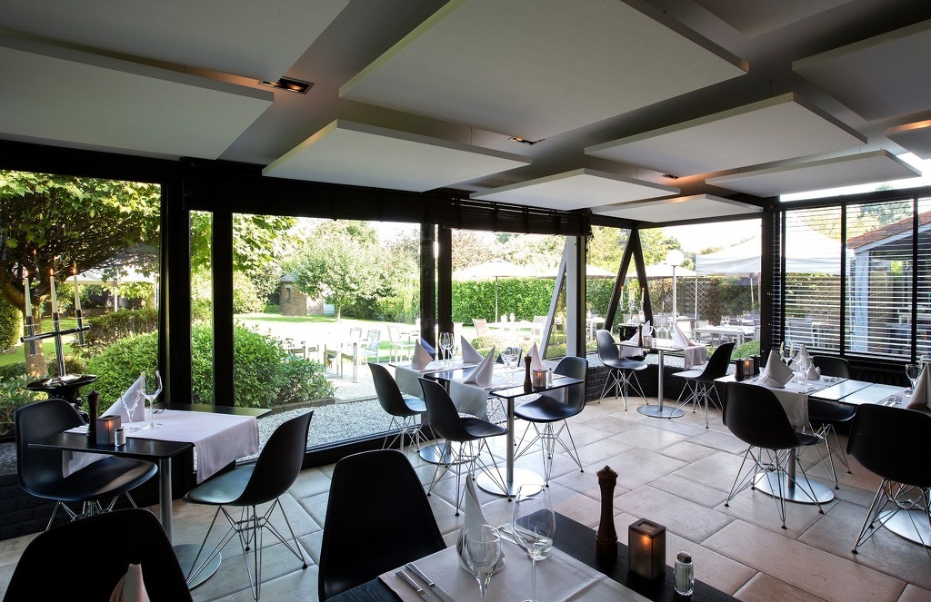 Eleganckie wnętrze restauracji z widokiem na zielony ogród, czarne krzesła Eames i nakryte stoły z białymi obrusami i serwetkami, widoczne duże okna z czarnymi ramami.