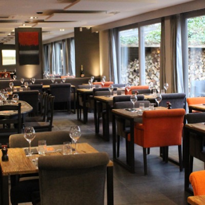 Wnętrze restauracji z drewnianymi stołami, szarymi krzesłami i pomarańczowymi akcentami, widok na stos drewna za oknem.