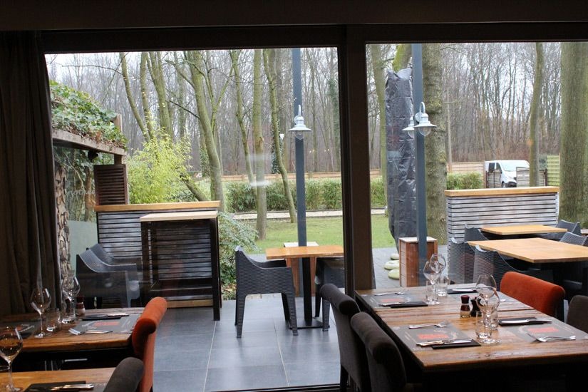 Zadaszony taras restauracji z widokiem na ogród, stoliki nakryte do posiłku, na zewnątrz widoczne drzewa i elementy małej architektury.