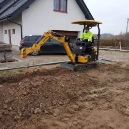 Żółta mini koparka NEX z operatorem w odblaskowej kamizelce, przygotowująca teren pod budowę, widoczny wykop, hałda ziemi i paleta z kostką brukową w tle domu z czarnym samochodem.
