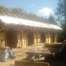 Wznoszenie drewnianej konstrukcji altany z widocznym szkieletem dachu pokrytym białą membraną, w tle drzewa i fragment dachu innego budynku.