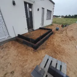 Przygotowanie podłoża pod taras z obrzeżami z kostki brukowej, widoczne szpilki wyznaczające poziom, piasek i paleta z kostką obok.