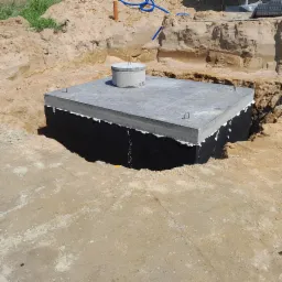 Wykonany, zaizolowany fundament z wystającymi prętami zbrojeniowymi, otoczony wykopem w piaszczystej ziemi.